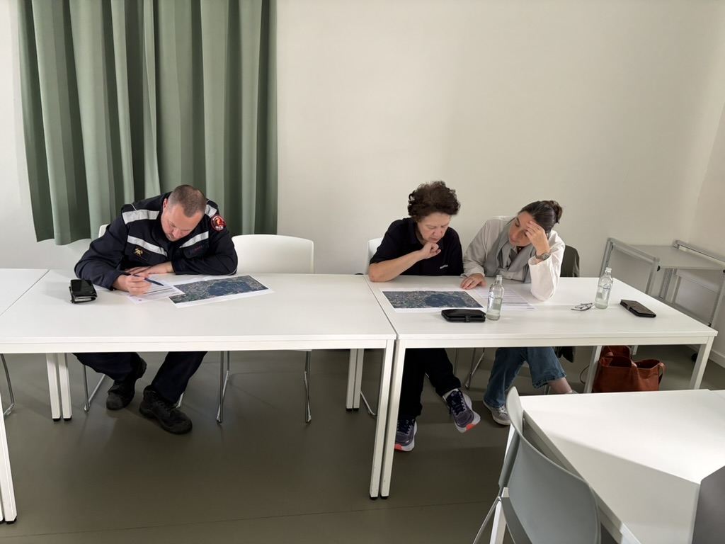 Een brandweerman en twee deelnemers bestuderen luchtfoto’s van de case aan een tafel